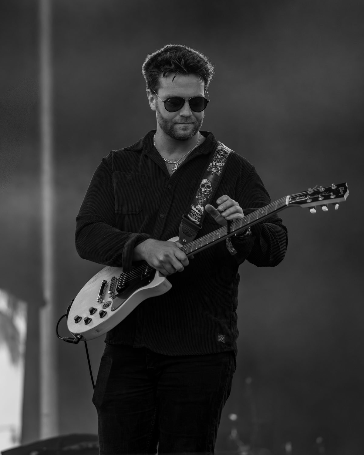 Black and white photo of Brian Lawless playing guitar on stage in a corduroy shirt and sunglasses
